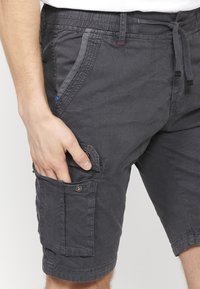 Handen vilar i sidofickan på mörkgrå cargoshorts med dragsko i midjan, som bärs av en person i en vit skjorta.