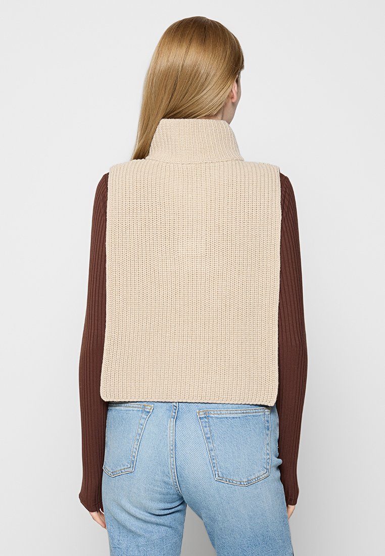 Frau mit glattem blonden Haar, die eine beigefarbene gestrickte ärmellose Weste über einem braunen Langarmshirt und hellblauen Jeans trägt, steht mit dem Rücken zur Kamera.