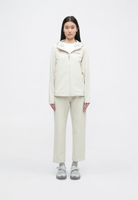 Veste beige avec capuche et fermeture éclair à l'avant, associée à un pantalon beige assorti. Le matériau semble lisse ; les chaussures sont grises avec une sangle.