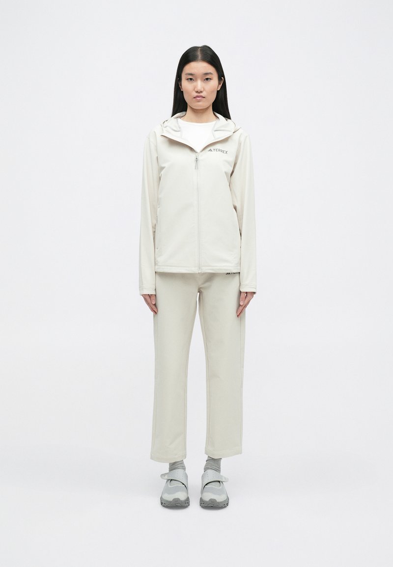 Veste beige avec capuche et fermeture éclair à l'avant, associée à un pantalon beige assorti. Le matériau semble lisse ; les chaussures sont grises avec une sangle.