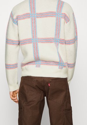 Personne portant un pull crème avec des motifs à carreaux bleu et rouge et un pantalon marron foncé vue de dos.