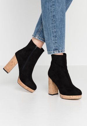 Zwarte suède enkelboots met een kurken blokhak, voorzien van een zijrits en een ronde neus, gedragen met enkelhoge blauwe jeans.