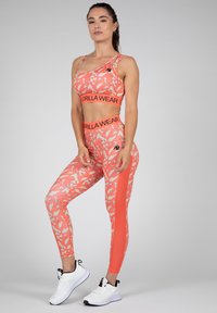 Koraal sportset bestaande uit een sportbeha met één schouder en hoge taille leggings met een lichtcrème abstract patroon. Witte sneakers maken de look compleet.