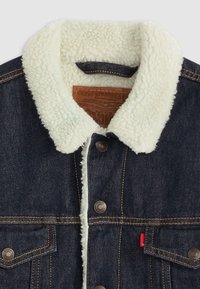 Chaqueta de mezclilla con un cuello de borreguito falso en color crema. Cierre de botones, costuras en color cobre y una etiqueta de cuero visible en la parte interna del cuello.