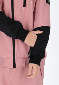 Pink sportjakke med sorte ærmer, ribbede manchetter og en lynlåslomme på venstre arm. Glat stof med en let glans.