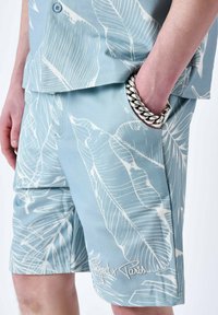 Camisa de manga corta azul claro y pantalones cortos a juego con un patrón de hojas blancas; los pantalones cortos cuentan con bolsillos laterales y un detalle de logo bordado.