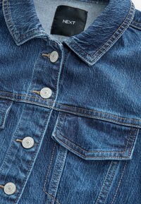 Denim jacket with a blue texture, featuring silver hardware buttons, two chest pockets, and visible stitching detail. Brand label at the collar.