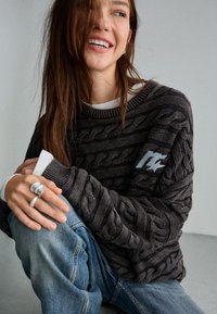 Gebreide trui met een donkergrijs kabelpatroon, ronde halslijn en gestreepte accenten. Gecombineerd met lichtblauwe jeans. Zilveren ring zichtbaar.