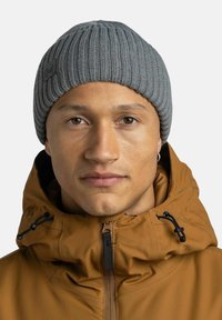 Gorro de punto gris con canalé y acabado texturizado. Lleva puesto una chaqueta con capucha marrón que cuenta con cremallera y ajustes de cordón.