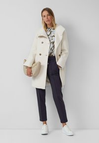 Frau in einem weißen strukturierten Mantel, floralem Blusenshirt, navyfarbener Hose, weißen Sneakers und einer beigen Handtasche mit goldener Kettenschlaufe.
