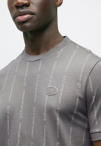 Άνδρας που φοράει γκρι μπλουζάκι EA7 Emporio Armani με κάθετα μοτίβα λογότυπου και στρογγυλό μεταλλικό σήμα με λογότυπο στο στήθος.