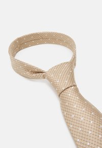 Cravate beige avec un motif texturé, présentant des rayures diagonales et de petits pois blancs. La cravate est nouée dans un style classique.
