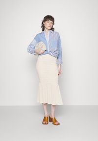 Camisa riscada azul e branca, saia bege com bainha rodada e sandálias de salto de madeira castanha. Bolsa de mão na mão, ambiente minimalista.