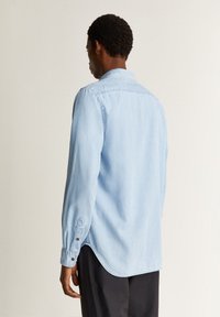 Lyseblå denimskjorte med lange ærmer, knap-ned krave og rund kant. Har to knapper på hver manchet og en glat overflade.