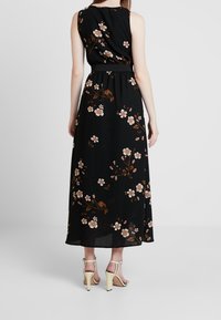 Nainen, jolla on yllään musta ilmanhihaisen kukallisen maksimekon ja vaaleat korkokengät, seisoo selkä kameraan päin yksinkertaista taustaa vasten.