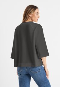 Vrouw die zich opzij draait, gekleed in een losse zwarte top met driekwartmouwen en een blauwe spijkerbroek, tegen een witte achtergrond.