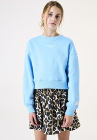 Sudadera cortada azul claro con mangas largas y texto blanco en el frente, combinada con una falda en capas con un patrón de estampado de leopardo.