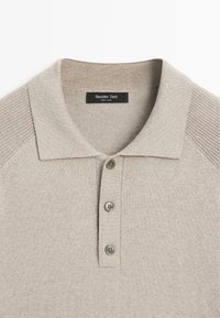 Polo beige con colletto a coste, tre bottoni grigi e dettagli strutturati sulle spalle. L'etichetta riporta "Massimo Dutti". Tessuto morbido e lavorato a maglia.