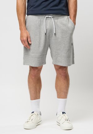 Man draagt grijze shorts met trekkoord, witte kniekousen en witte sneakers, staat met handen in de zakken.