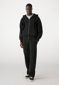 Zwarte rits-hoodie met een gevoerde capuchon, geribbelde manchetten en voorzakken, gecombineerd met losse zwarte broek en zwarte sneakers.