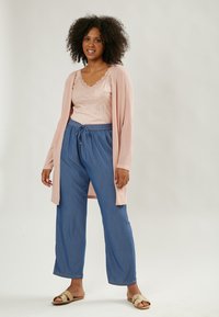 Cardigan rosa claro, top de tirantes rosa brillante, pantalones azules holgados y sandalias beige de deslizar, mostrando un atuendo cómodo y casual.