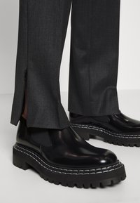 Bottes Chelsea épaisses en cuir noir avec semelles crantées épaisses et coutures blanches, portées avec un pantalon gris foncé présentant des fentes latérales et des fermetures éclair.