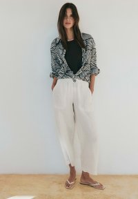 Woman standing against white wall wearing black top, patterned black-and-white shirt knotted at waist, white wide-leg pants, and white sandals.