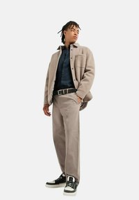 Giacca in pile beige sopra una camicia a bottoni blu navy, abbinata a pantaloni beige chiaro e sneaker nere con dettagli bianchi.