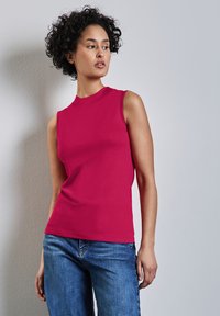 Sovittava, hengettömän toppi kirkkaan vaaleanpunaisesta puuvillasta, jossa on korkea kaula-aukko; käytetty vaaleansinisien farkkujen kanssa, yksinkertaisen taustan edessä.