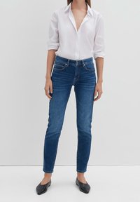 Blue denim jeans with a slim fit, featuring a mid-rise waist, five pockets, and subtle stitching details. Worn with a white button-up shirt.