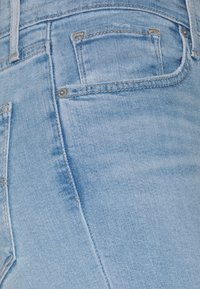 Gros plan sur un jean en denim bleu clair montrant la poche avant, la passants de ceinture et les détails visibles des coutures.