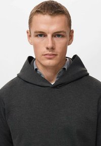 Donkergrijze hoodie met een grote, zachte capuchon. Gemaakt van gladde stof en heeft een minimalistisch ontwerp. Gelaagd over een lichtblauw shirt.