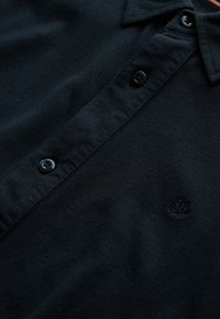 Navyblaue Polohemden aus strukturiertem Stoff, mit einer Dreiknopf-Leiste, schwarzen Knöpfen und einem kleinen gestickten Logo auf der Brust.