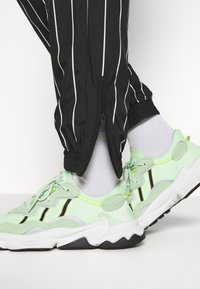 Vaaleanvihreät verkkokengät mustilla yksityiskohdilla, kuvioidulla pohjalla ja nauhakiinnityksellä, yhdistetty mustiin raidallisiin vetoketjullisiin verryttelyhousuihin.