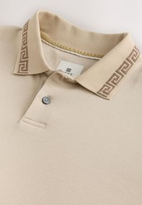 Beige Poloshirt aus glattem Stoff, mit einem Kragen und einem gemusterten braunen Akzent sowie einem einzelnen grauen Knopf an der Knopfleiste.