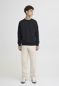 Melns kokvilnas džemperis ar ribveida apkakli, kombinēts ar gaiši bēšām biksēm. Modelis nēsā baltas un pelēkas sporta kurpes ar melnām detaļām.