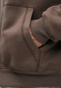 Hnedá mikina s mäkkou textúrou, s predným vreckom a rebrovanými manžetami. Stehy majú o niečo tmavší odtieň hnedej farby.