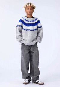 Niño con rastas rubias claras que lleva un suéter gris con patrones azules y blancos, pantalones vaqueros grises holgados y zapatillas blancas con rayas negras.