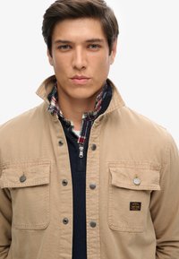 Beige utilityjack met twee voorzakken, zilveren hardware en een geruite kraag. Draagt over een marineblauw jack met ritssluiting van een gladde textuur.