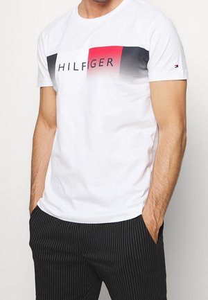 Man in white Tommy Hilfiger T-shirt with red and black gradient logo, wearing black pinstripe trousers, hands in pockets, against plain background.