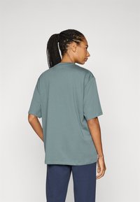 Prevelik t-shirt v potopljeni teal barvi, kratki rokavi, krožni izrez, izdelan iz mehke tkanine, prikazan s hrbta z zapletenimi lasmi.