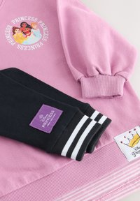 Jersey rosa con mangas negras. Presenta un gráfico de personaje y una etiqueta morada. Puños acanalados con rayas blancas y un parche con diseño de corona.