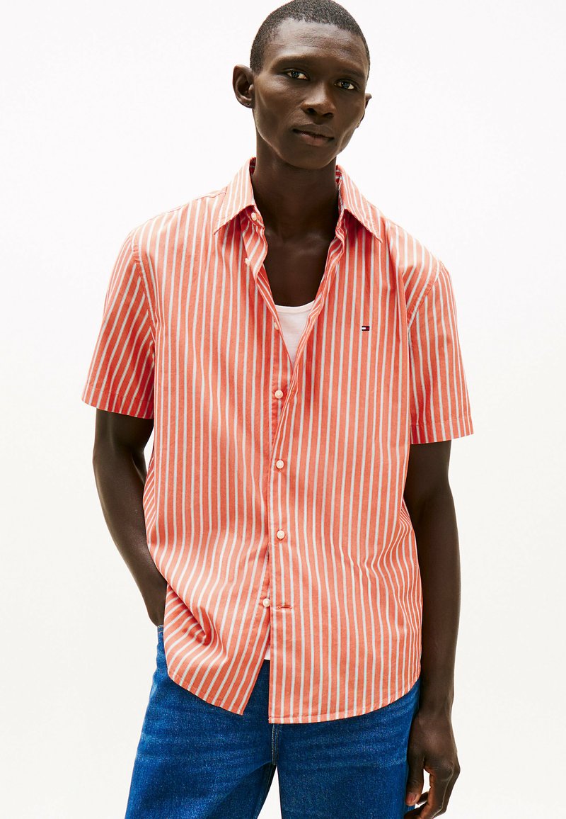 Camisa de botones de manga corta a rayas coral y blancas. Con cuello, un logo en el pecho, fabricada en tejido ligero, combinada con unos jeans de mezclilla.