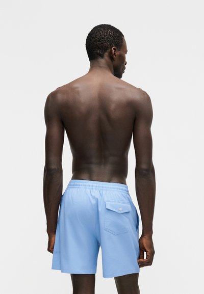 Mees seisab paljajalu, vaadatuna tagant, kandes helesiniseid ujumispükse tagatasku ja elastse vööga.