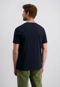 Man met bruin haar die een eenvoudig zwart t-shirt met korte mouwen en groene corduroybroeken draagt, met zijn rug naar de camera gekeerd tegen een witte achtergrond.