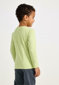 Lichtgroen T-shirt met lange mouwen, een ronde hals, gladde stof en een losse pasvorm. Gecombineerd met een blauw broek, gezien vanaf de achterkant.