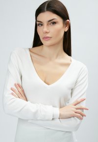Maglione bianco a maniche lunghe con scollatura a V e design aderente. Texture morbida e polsini a costine. Il modello è in piedi con le braccia incrociate, su uno sfondo neutro.