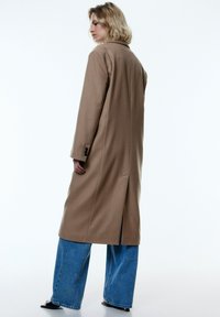 Beige wollen jas met een geknipt kraag, enkele achteropening en knoopmanchetten, gecombineerd met blauwe denim jeans met brede pijpen.