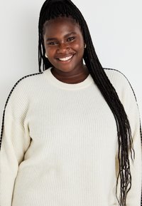 Κρέμ μαχαιρωτή ζακέτα με υφή και μαύρες λεπτομέρειες στο πλάι, διαθέτοντας στρογγυλή λαιμόκοψη και ελαφρώς φαρδιά εφαρμογή.