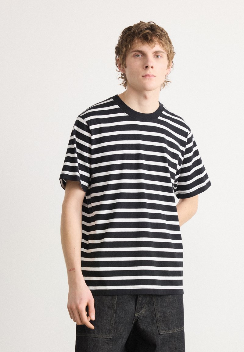 Tricou cu dungi alb-negru, cu decolteu rotund și mâneci scurte. Țesătura pare moale; croiul este lejer. Poartă-se cu pantaloni scurți din denim închisi la culoare.